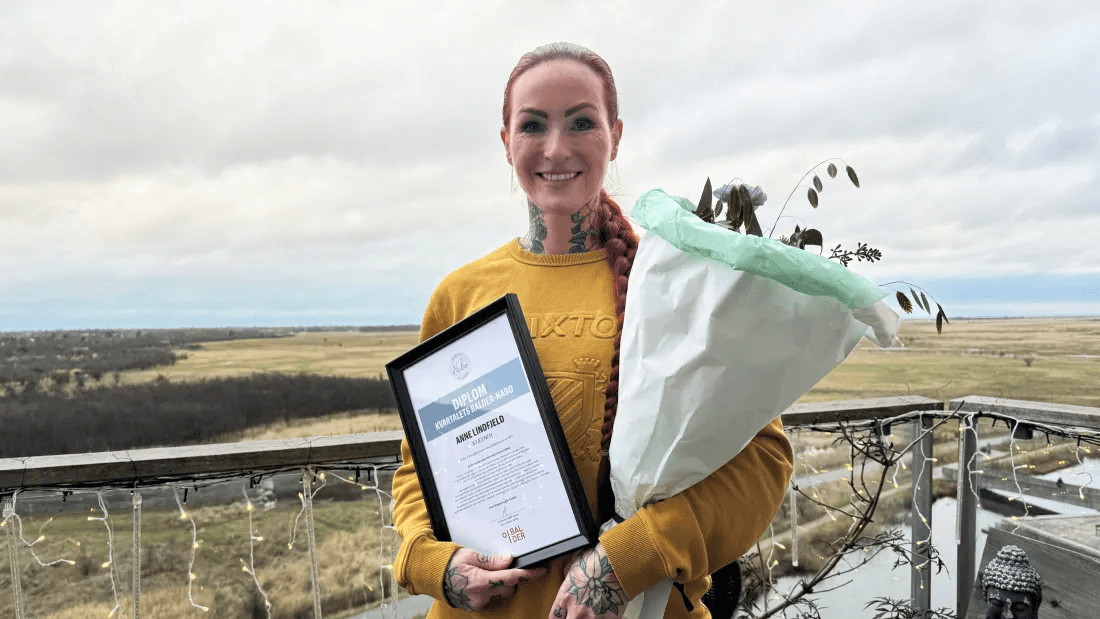 Anne Lindfjeld stands on her balcony with flowers and a certificate for Balder Neighbour of the Quarter.