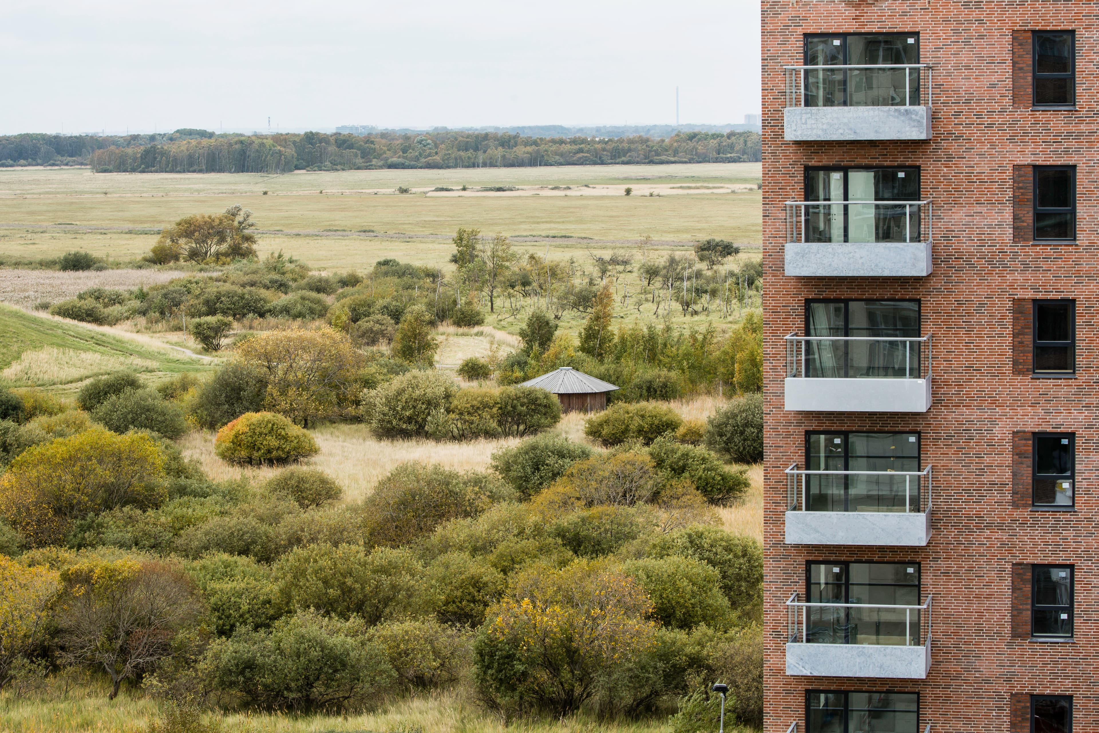 Ejendommen Elsehus eksteriørfoto med en murstensfacade med altaner til højre og udsigt over et grønt naturområde med buske, træer og åbne marker.