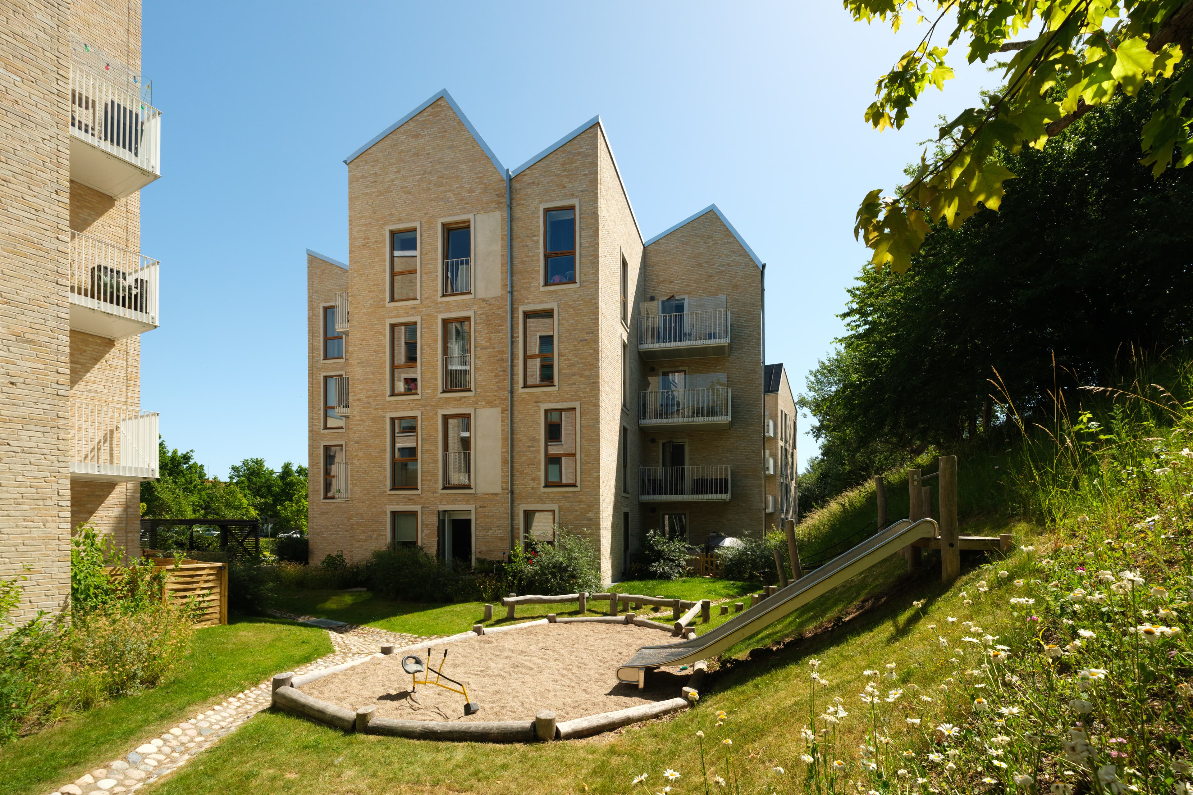 Green area by the property’s playground with a slide. The property is located in Hundige, and the façade of StrandbyHuse is visible with its roof structure built up in multiple peaks.