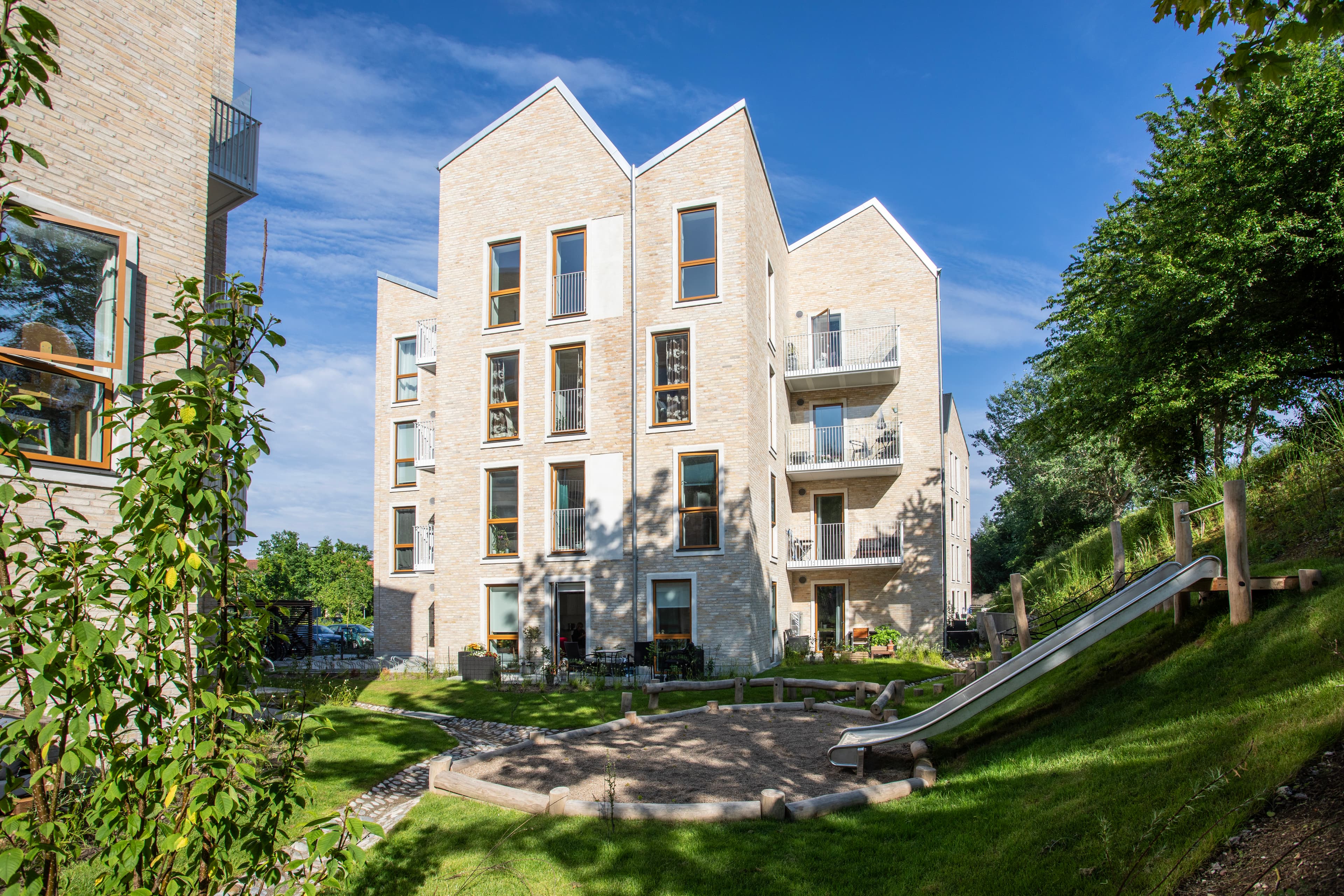 Grønt område ved ejendommens legeplads med rutsjebane. Ejendommen ligger i Hundige, og Strandby Huses facade ses med sin tagkonstruktion, som er opbygget i flere spidser.